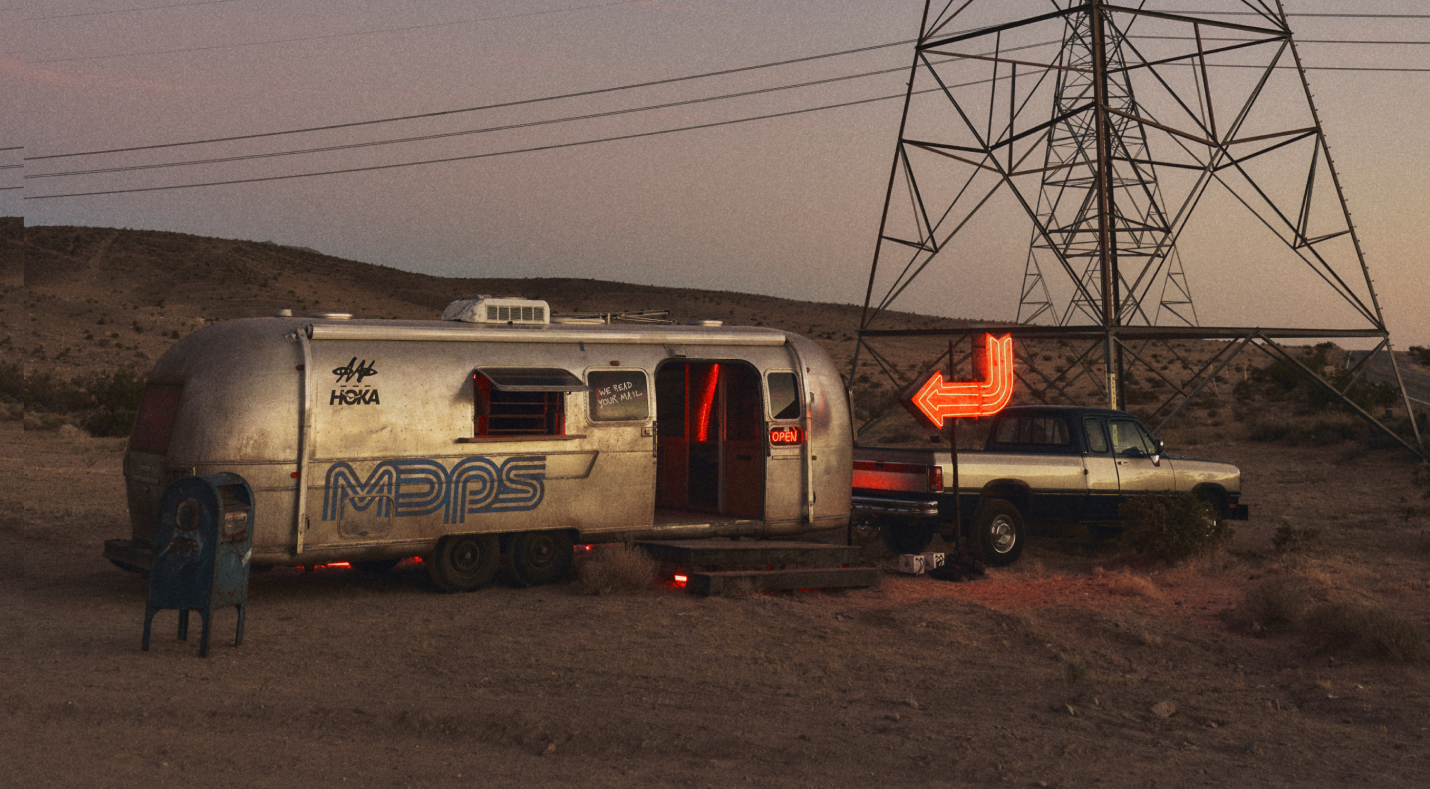 A wide shot of a silver vintage trailer converted into a postal station in the desert. It features the "MDPS" logo, a HOKA logo, and a neon orange arrow sign pointing toward it, situated under large power lines at dusk.