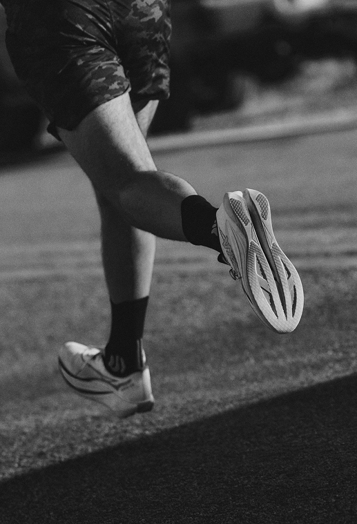 A black and white action shot of a runner's legs from behind, showing the detailed tread of a HOKA running shoe mid-stride.