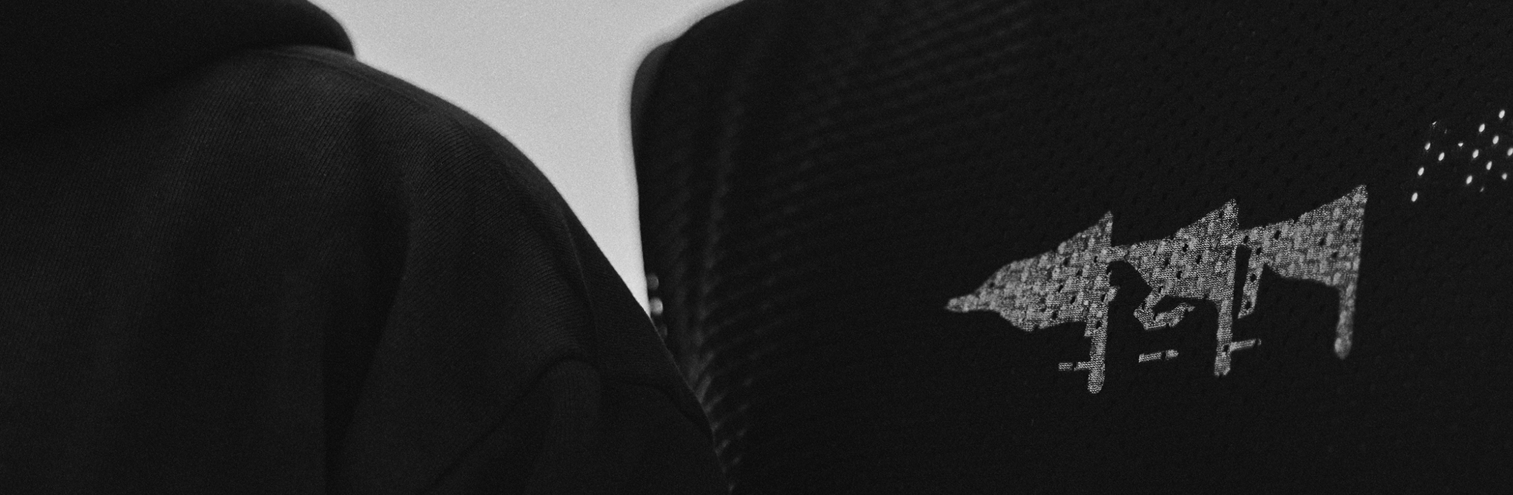 A close-up, black and white shot of a person's shoulder wearing a dark mesh athletic jersey with a stylized, textured graphic logo.