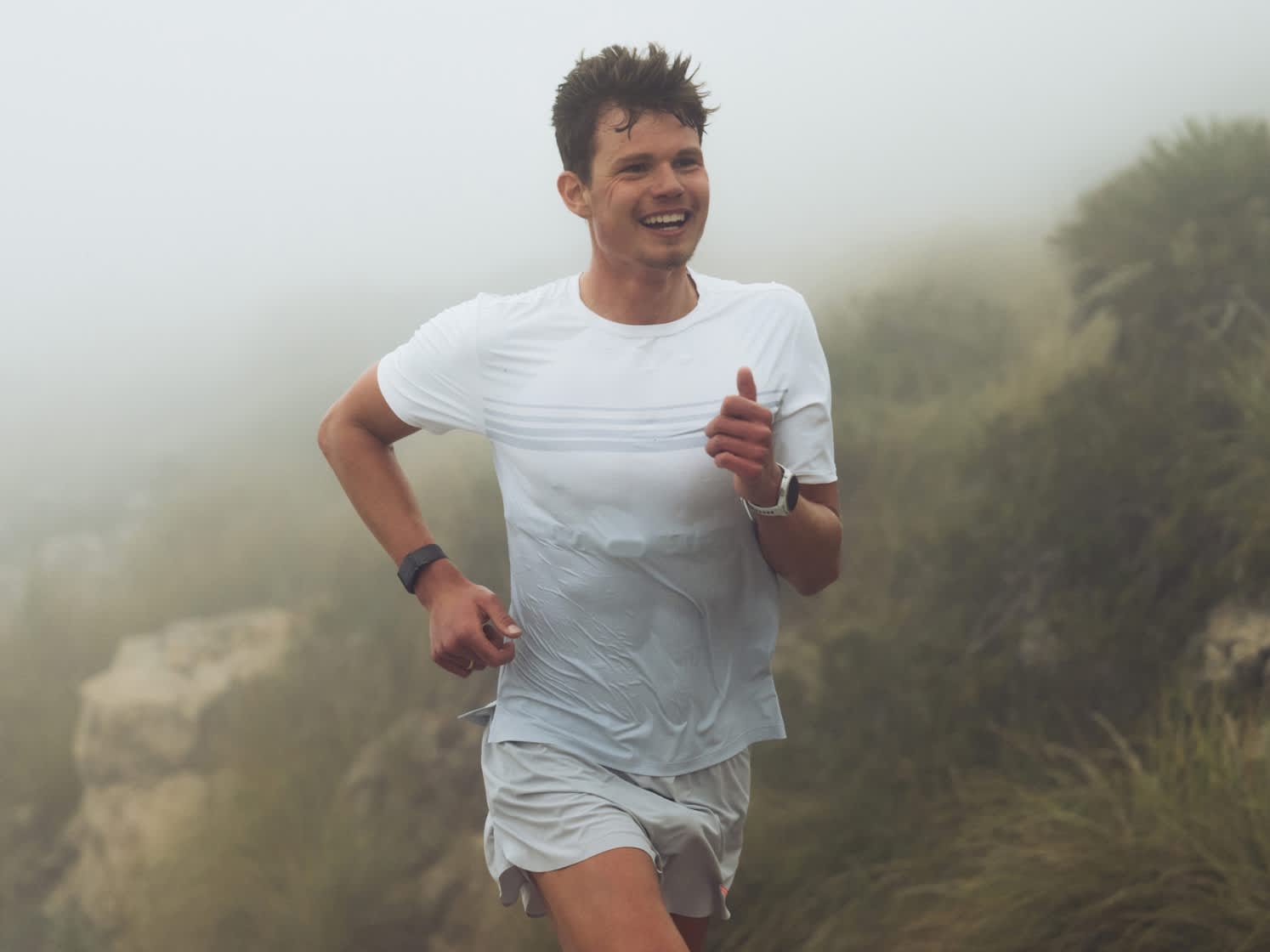 Smiling person running in white shirt and grey shorts through a misty landscape.