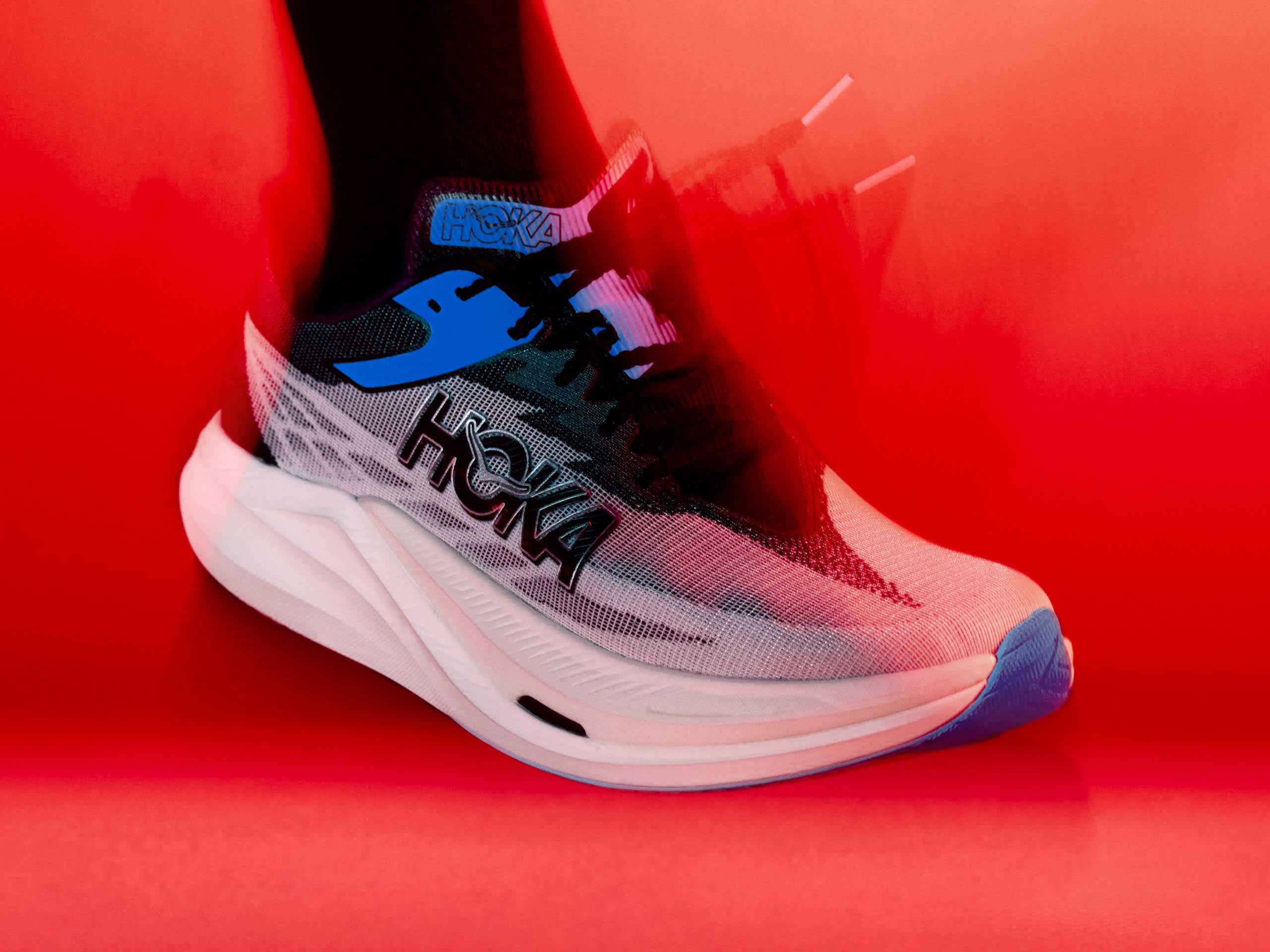 White and blue Hoka running shoe on a person's foot, against a vibrant red background. 