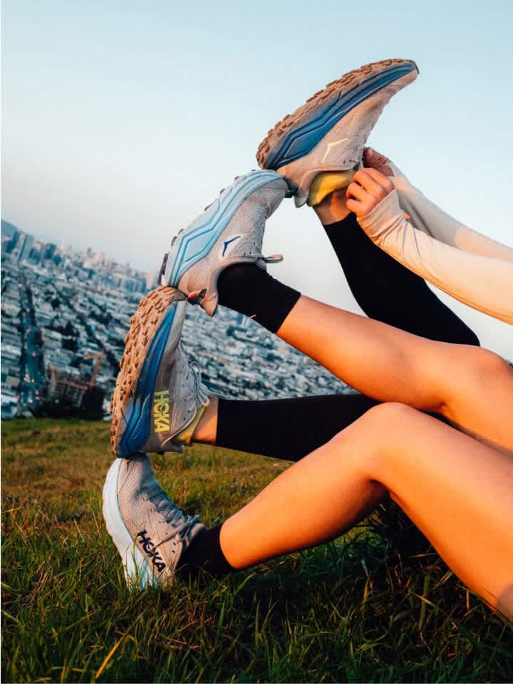 Intertwined legs with Hoka trail shoes on grass overlooking a city at sunset.