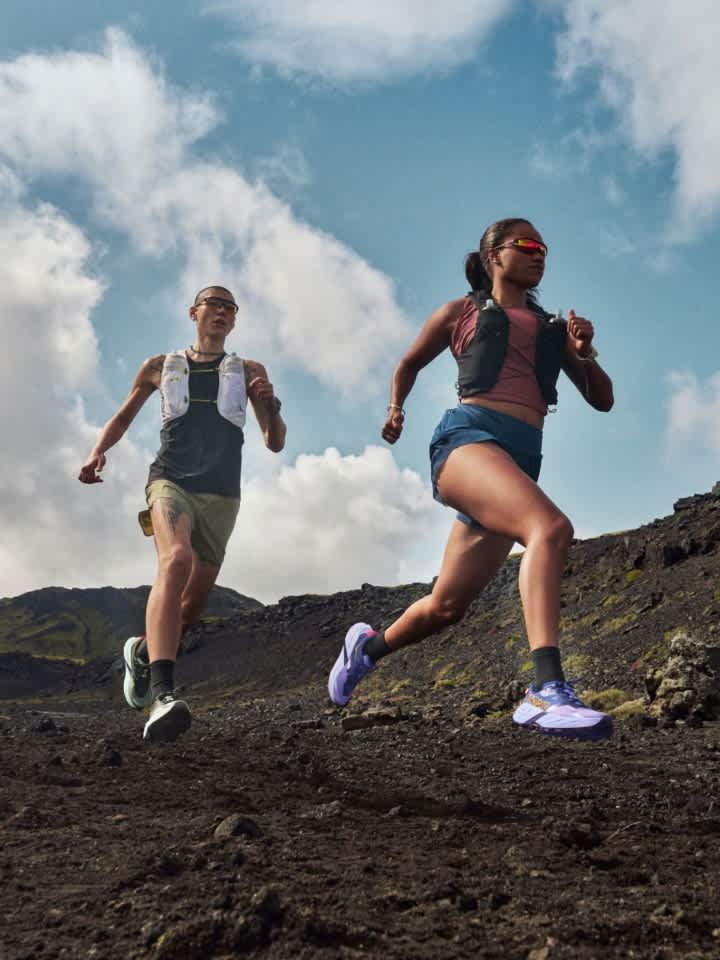 Two individuals trail running on dark, rocky terrain under a cloudy sky.