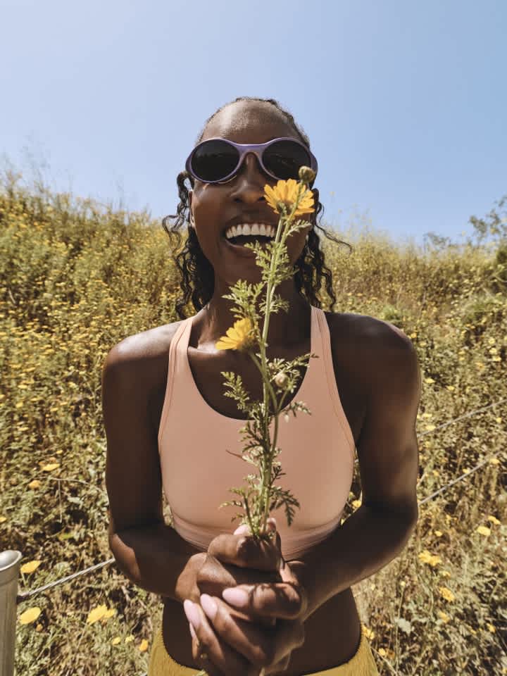 Smiling person in sunglasses holding yellow flowers in a field under blue sky.