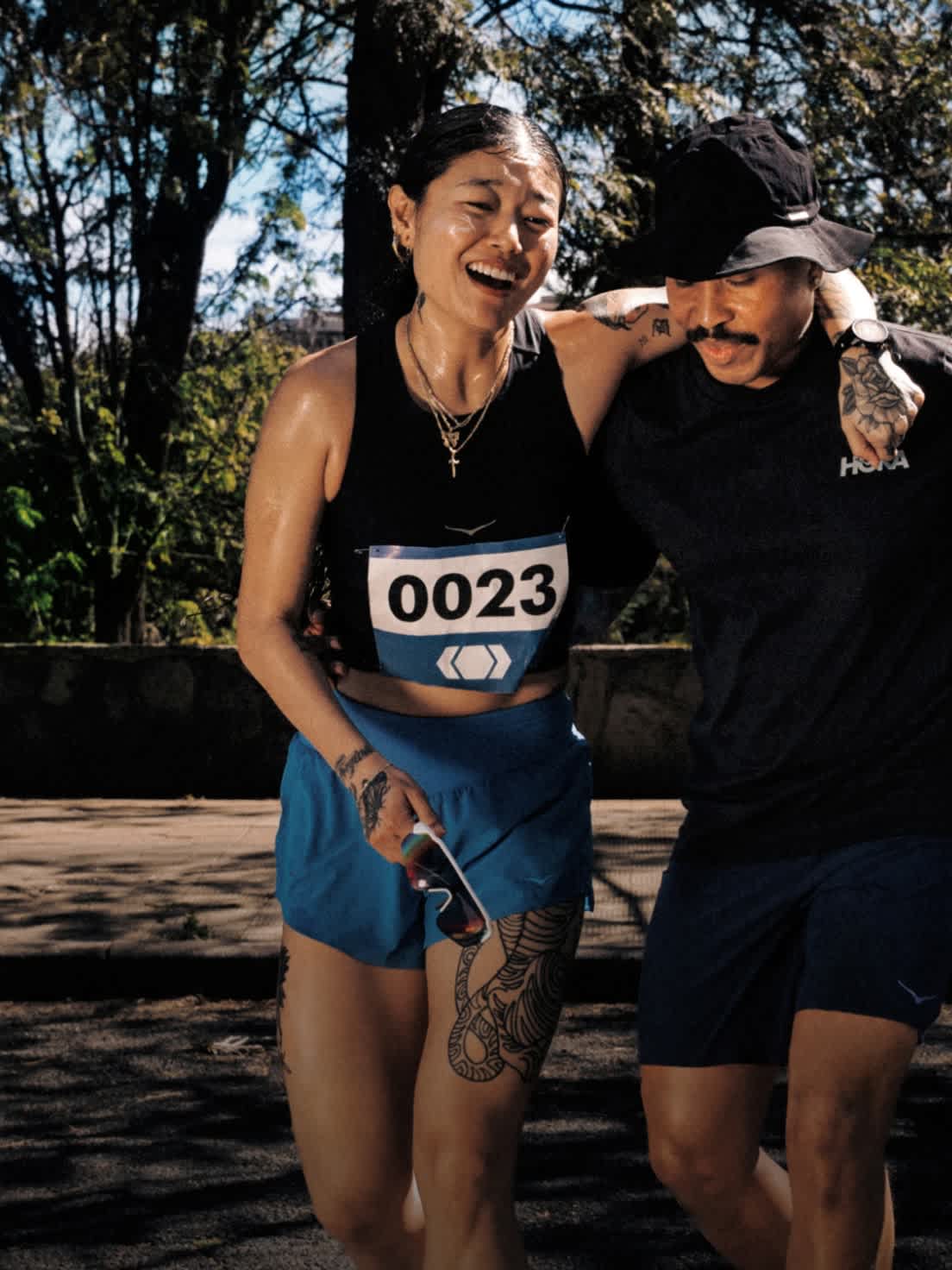 Sweating runner with bib 0023 smiles, supported by companion after race outdoors.