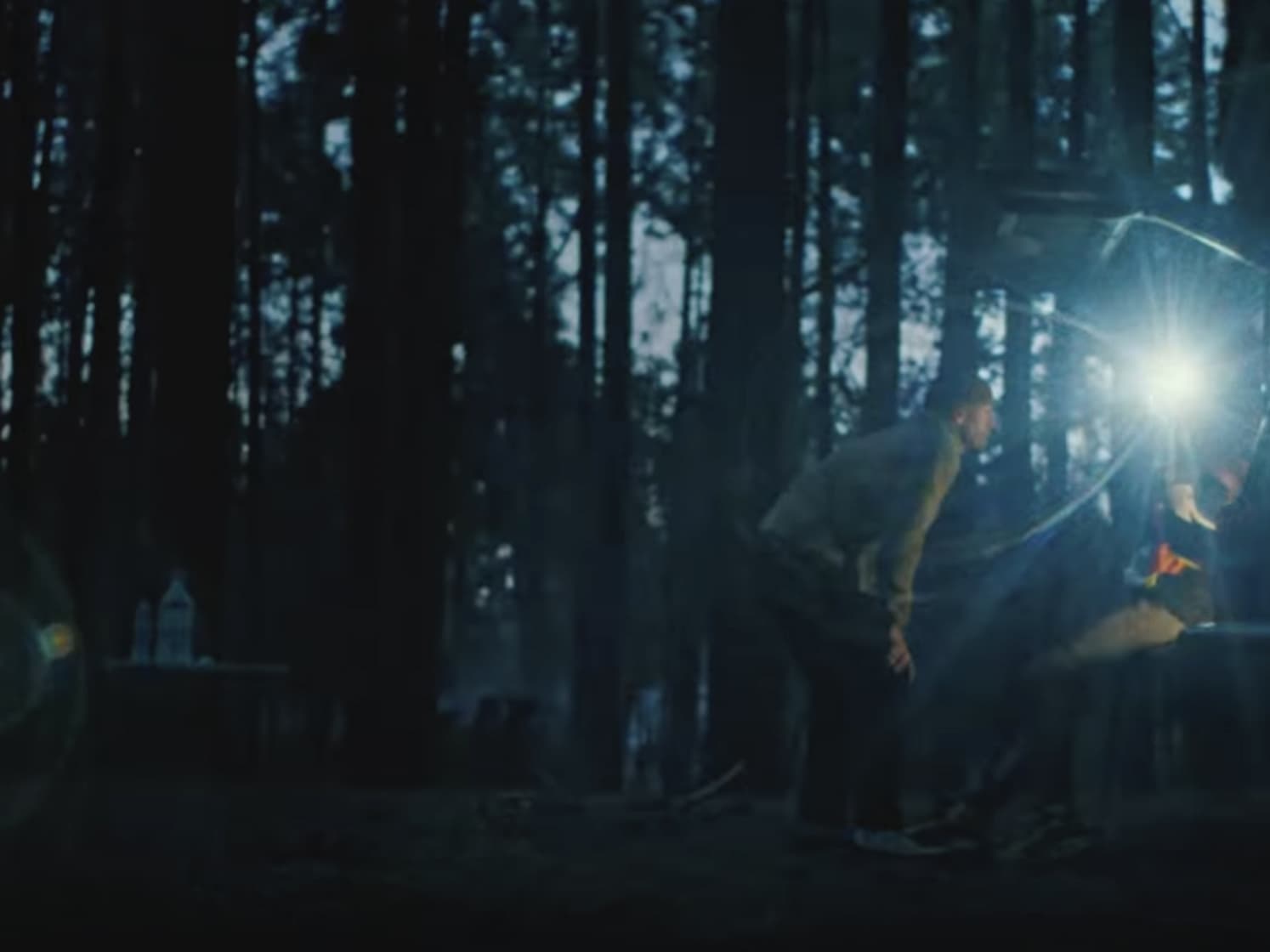 Dark forest, person bent over, illuminated by bright light; bottles on table left.