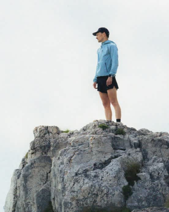 Runner in blue jacket and cap on rocky peak, looking left, against a bright sky.