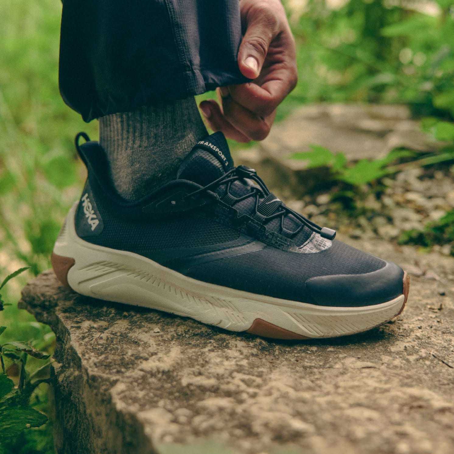 Primo piano del piede di una persona con scarpa Hoka scura su un sentiero di pietre tra foglie verdi.