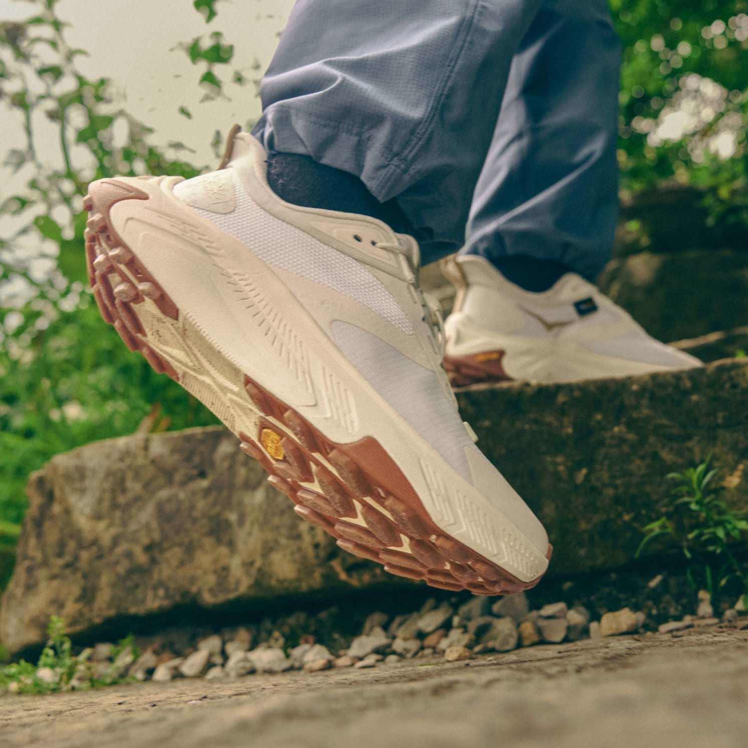 Persona con sneakers color panna con suola marrone che calpesta una pietra all’aperto.