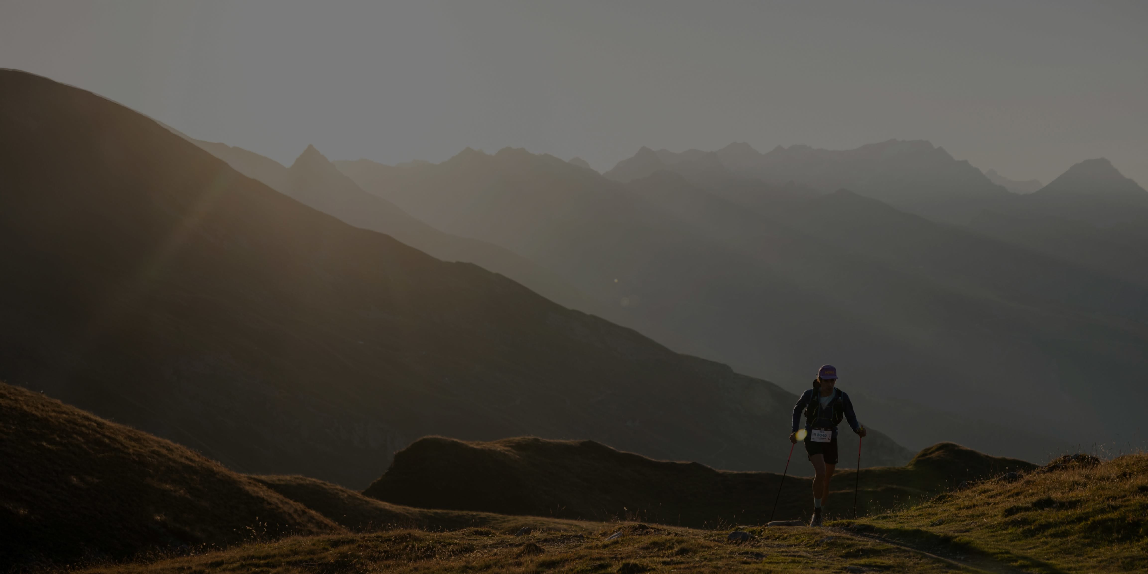 Person trail running with poles on a mountain path at sunset with layered peaks.