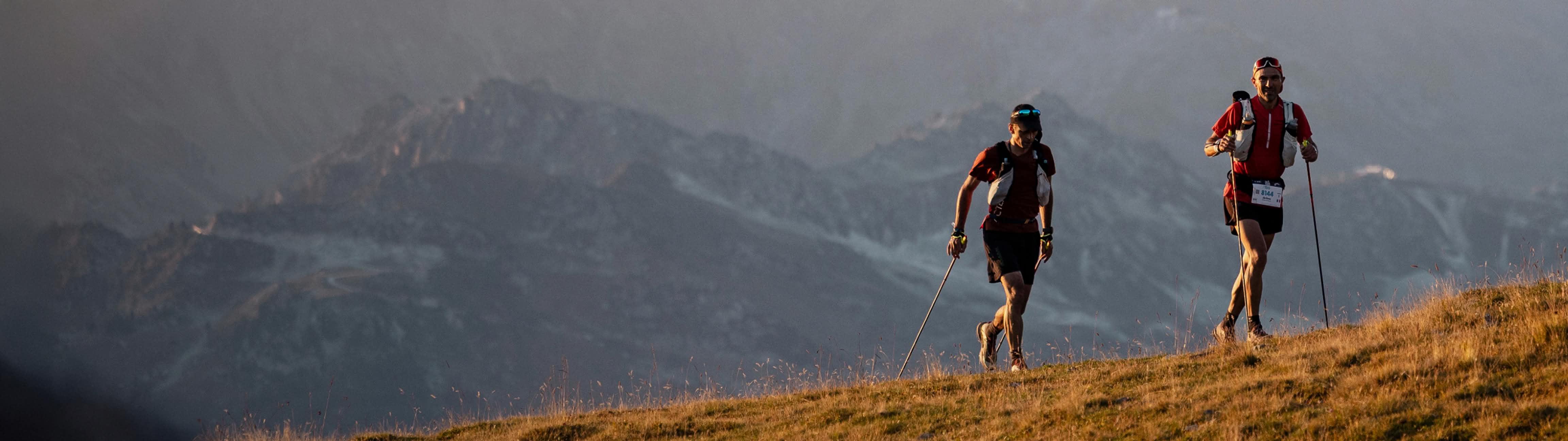 Two athletes trail running uphill with poles in golden light, misty mountains behind.
