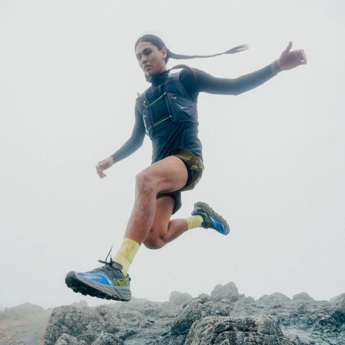 Athlete mid-jump on rocky terrain in Hoka trail shoes, long braids flying.
