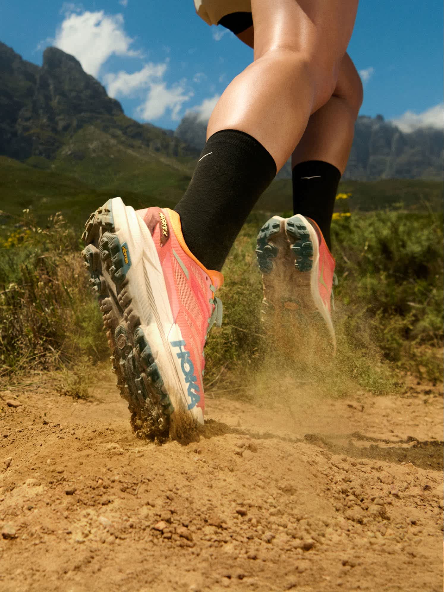Person running on a trail, HOKA shoes kicking dust, mountains in background.