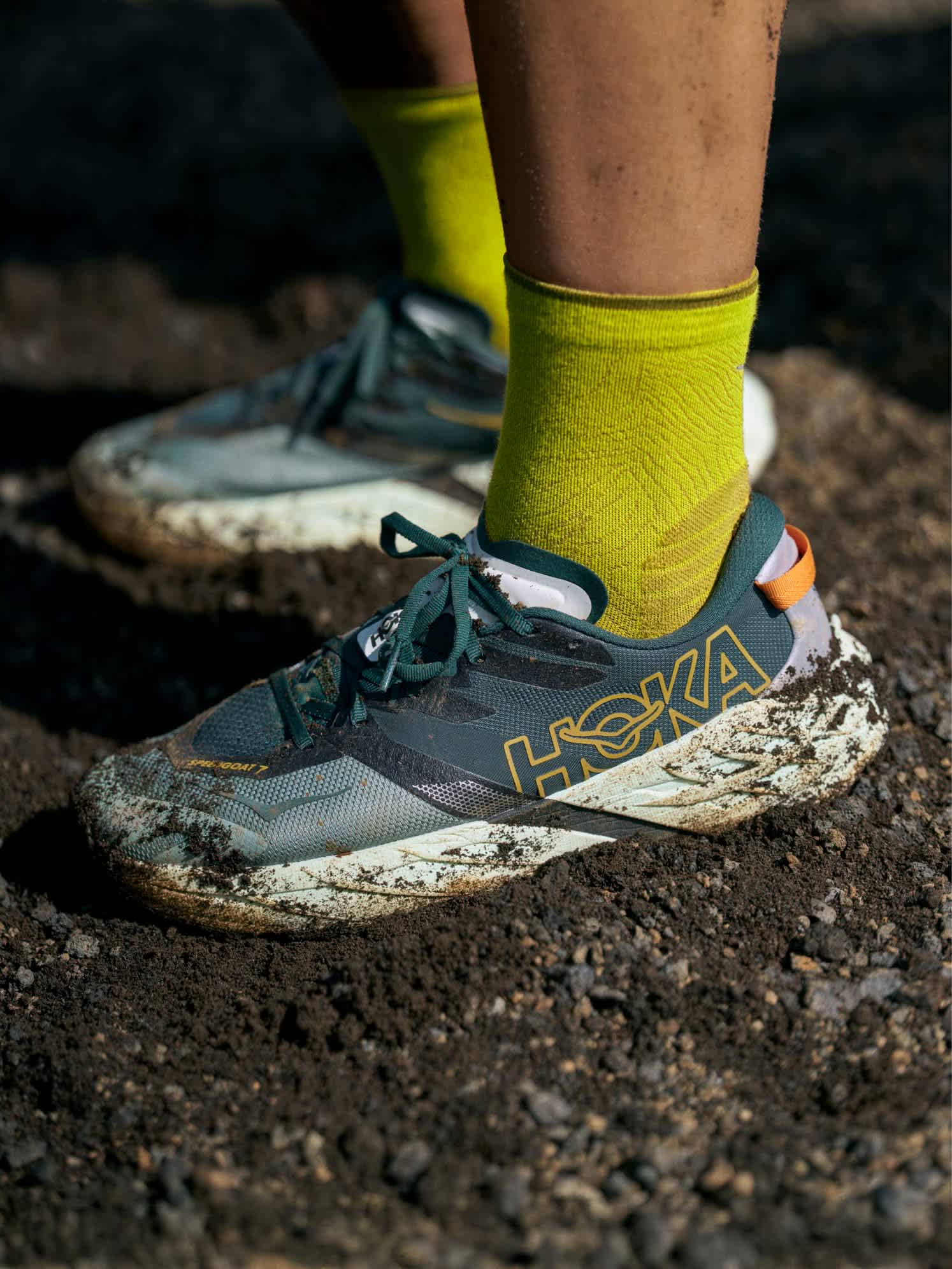 Close-up of muddy dark green Hoka Speedgoat 7 trail shoe with yellow socks on dirt.