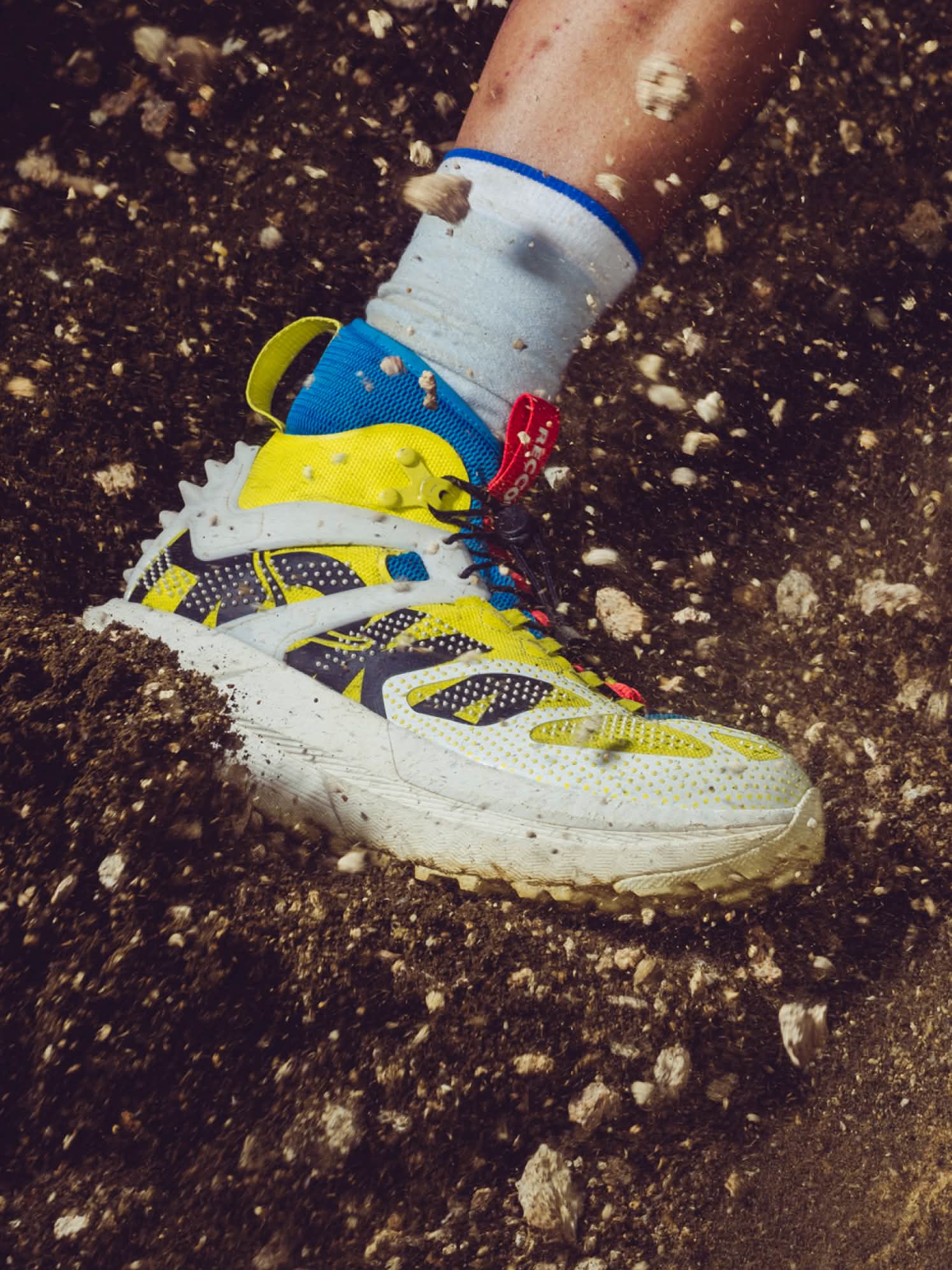 Colorful trail shoe, yellow and blue, kicking up dirt and rocks vigorously.