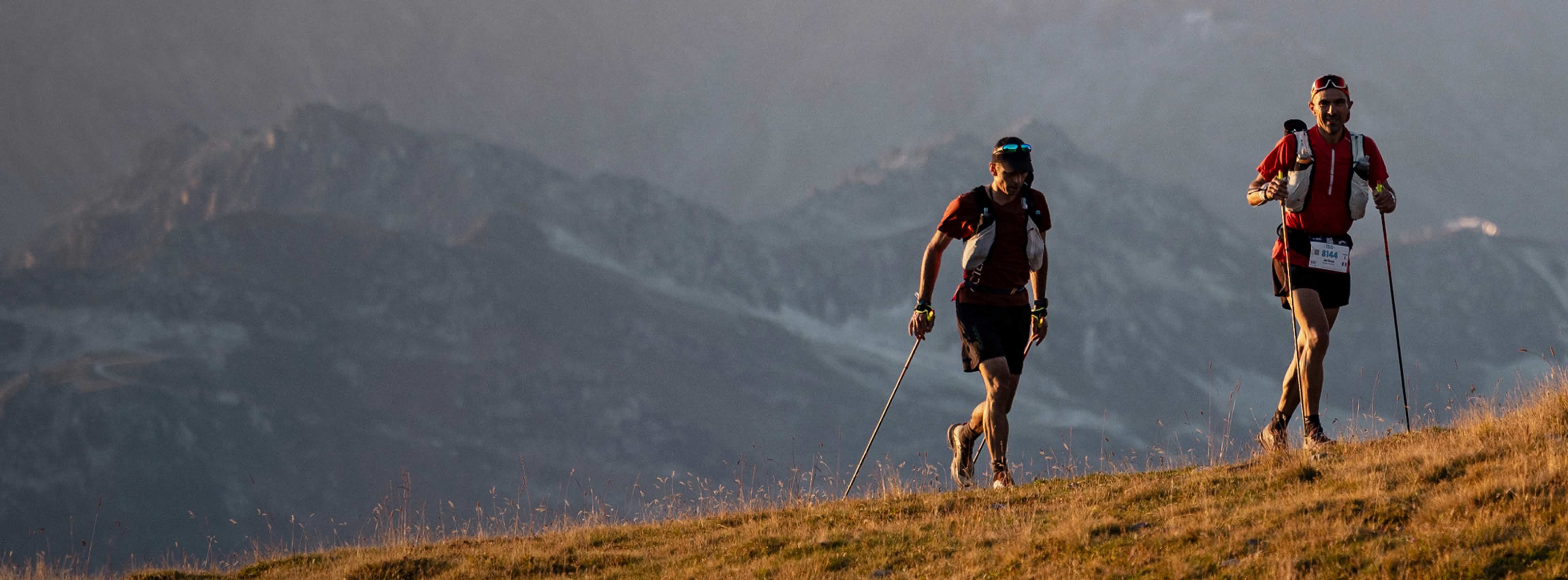 Two athletes run on a rocky path with distant mountains under a warm sky at dusk.