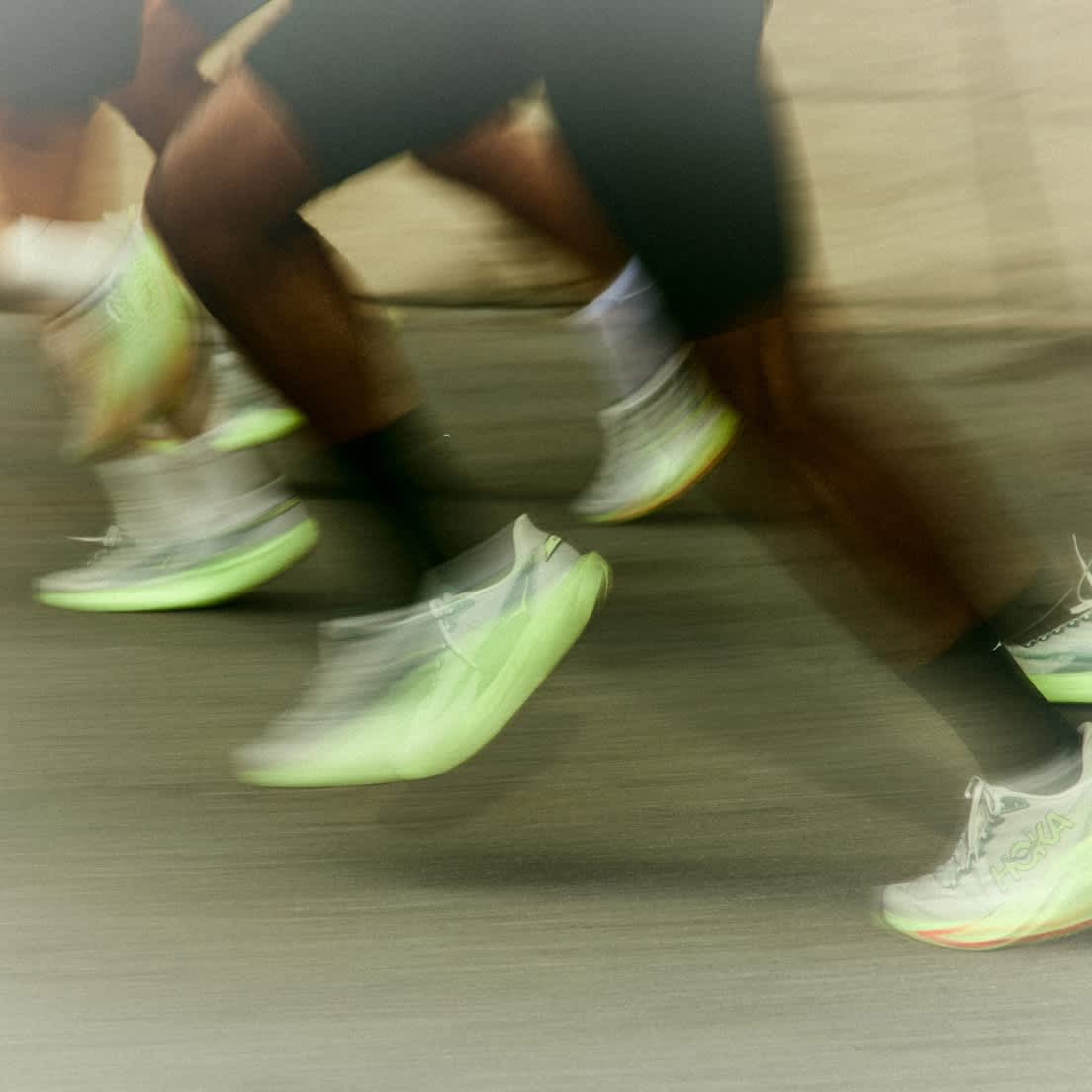 Blurred image of multiple runners' legs and bright green HOKA shoes in motion.