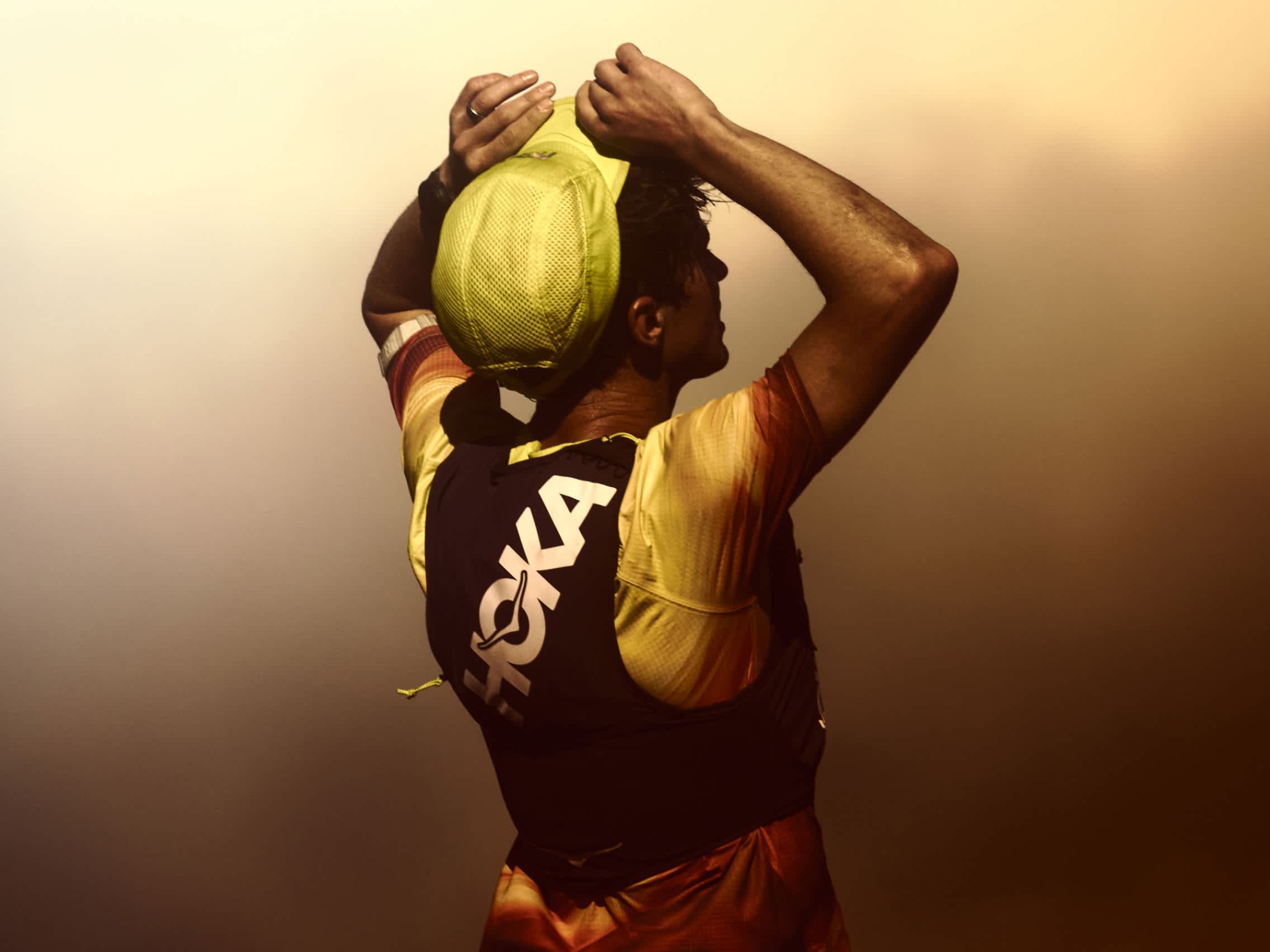 Runner from behind adjusting neon green cap, wearing HOKA vest, warm gradient light
