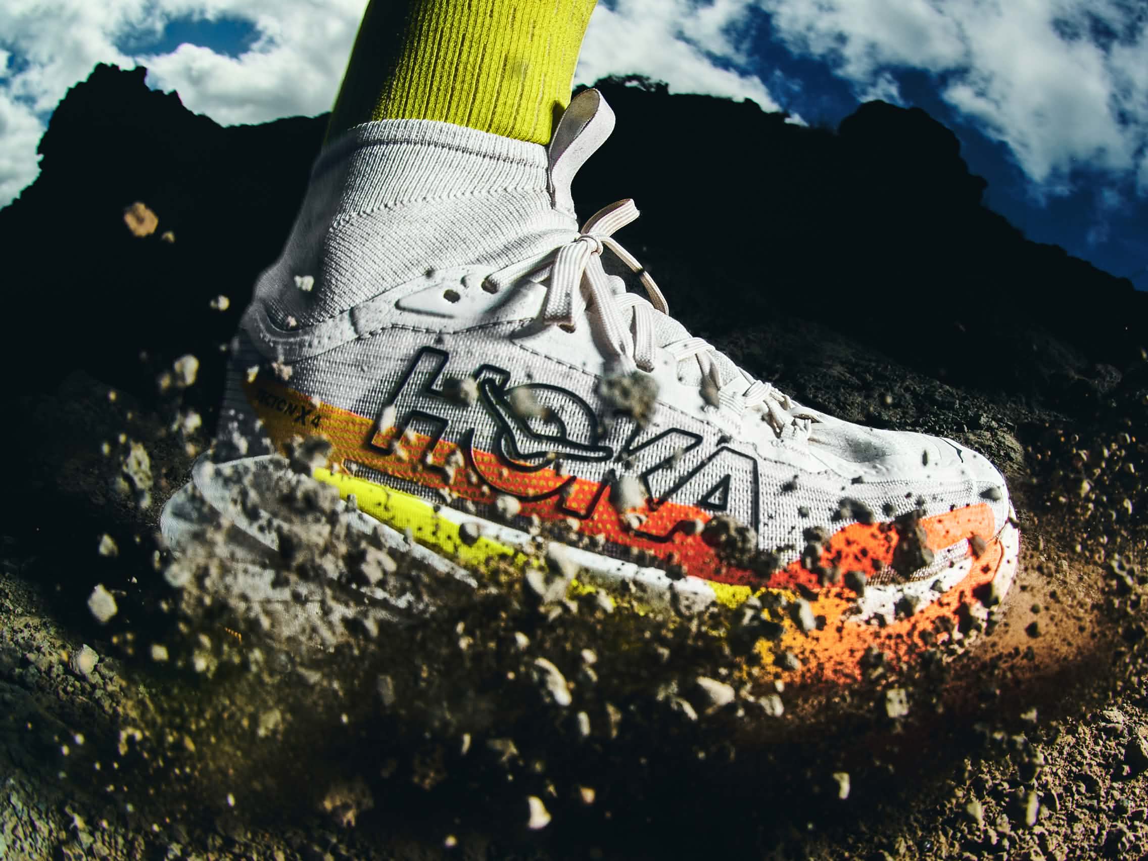 White Hoka trail running shoe kicking up dirt, with mountains and cloudy sky.