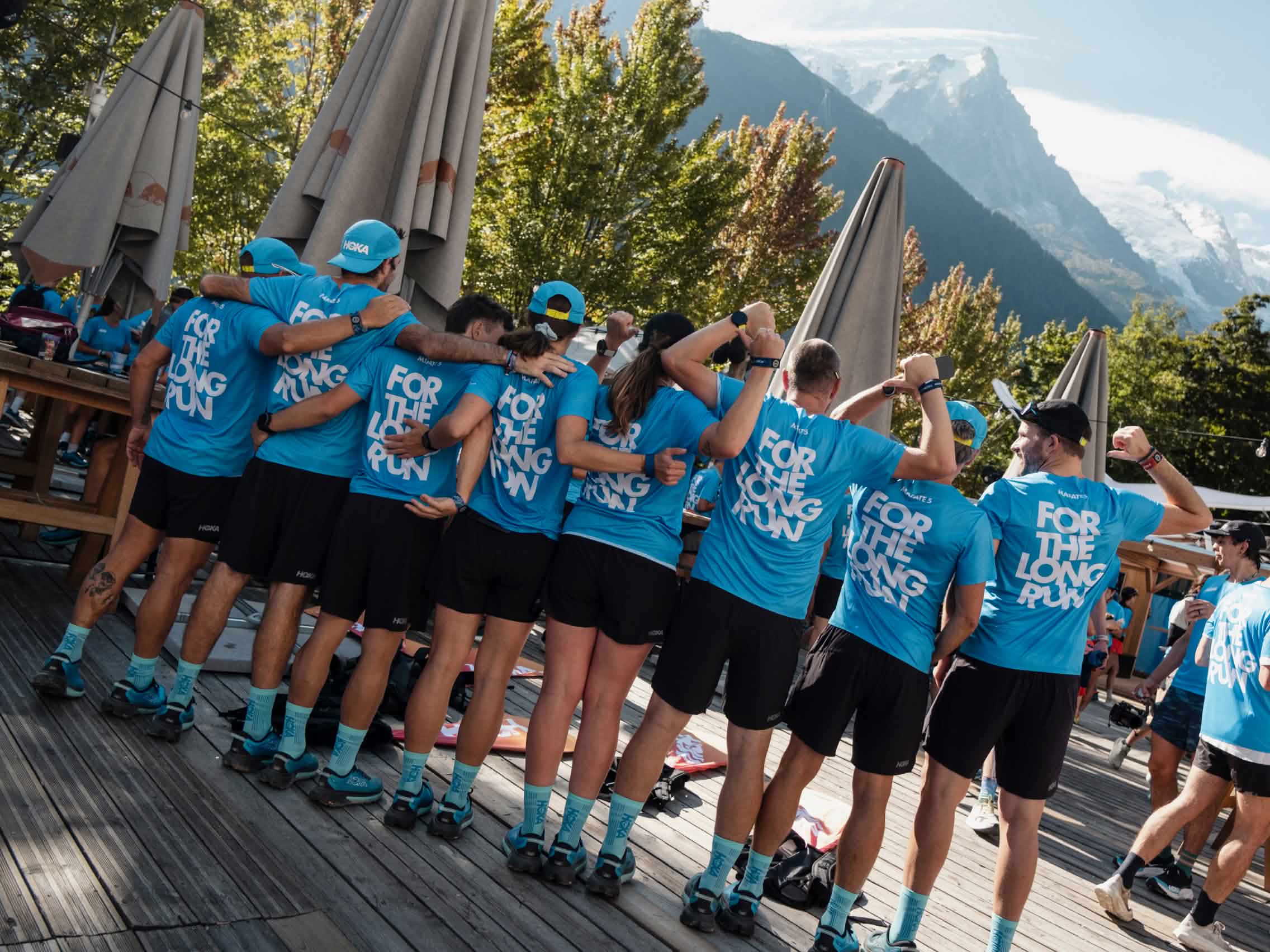 Group in blue HOKA shirts and shorts outdoors, some flexing, against mountains.