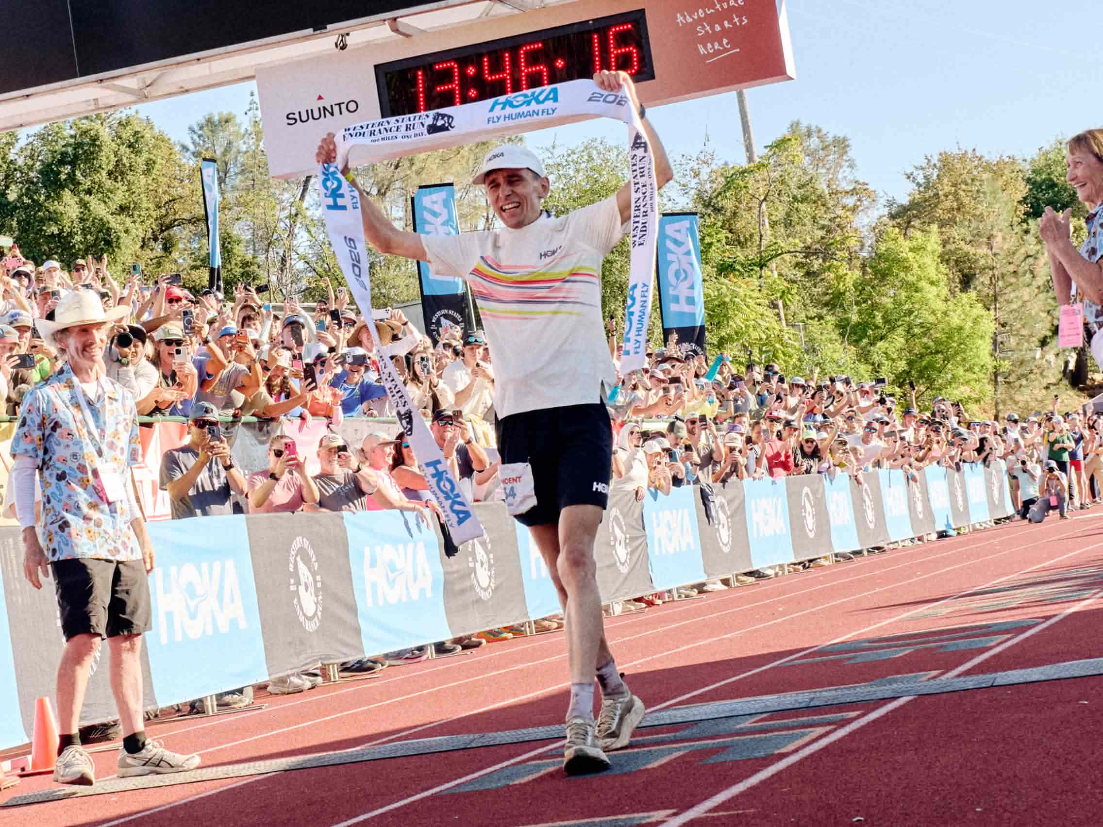 Person at finish line with banner, clock reads 13:46:16, surrounded by cheering