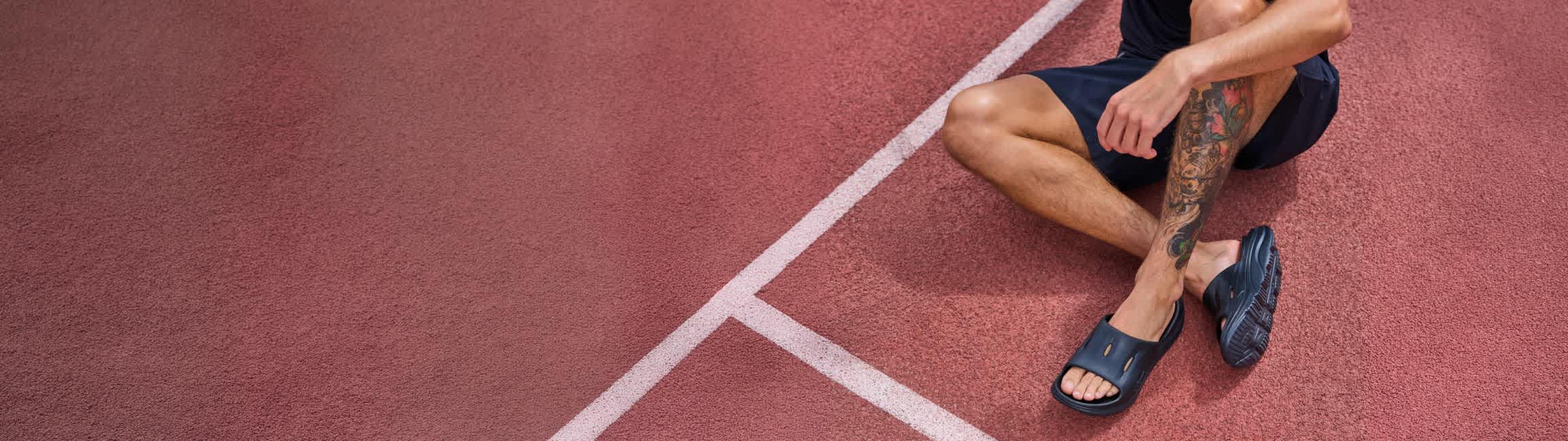 A runner relaxes on the track wearing HOKA Recovery slides