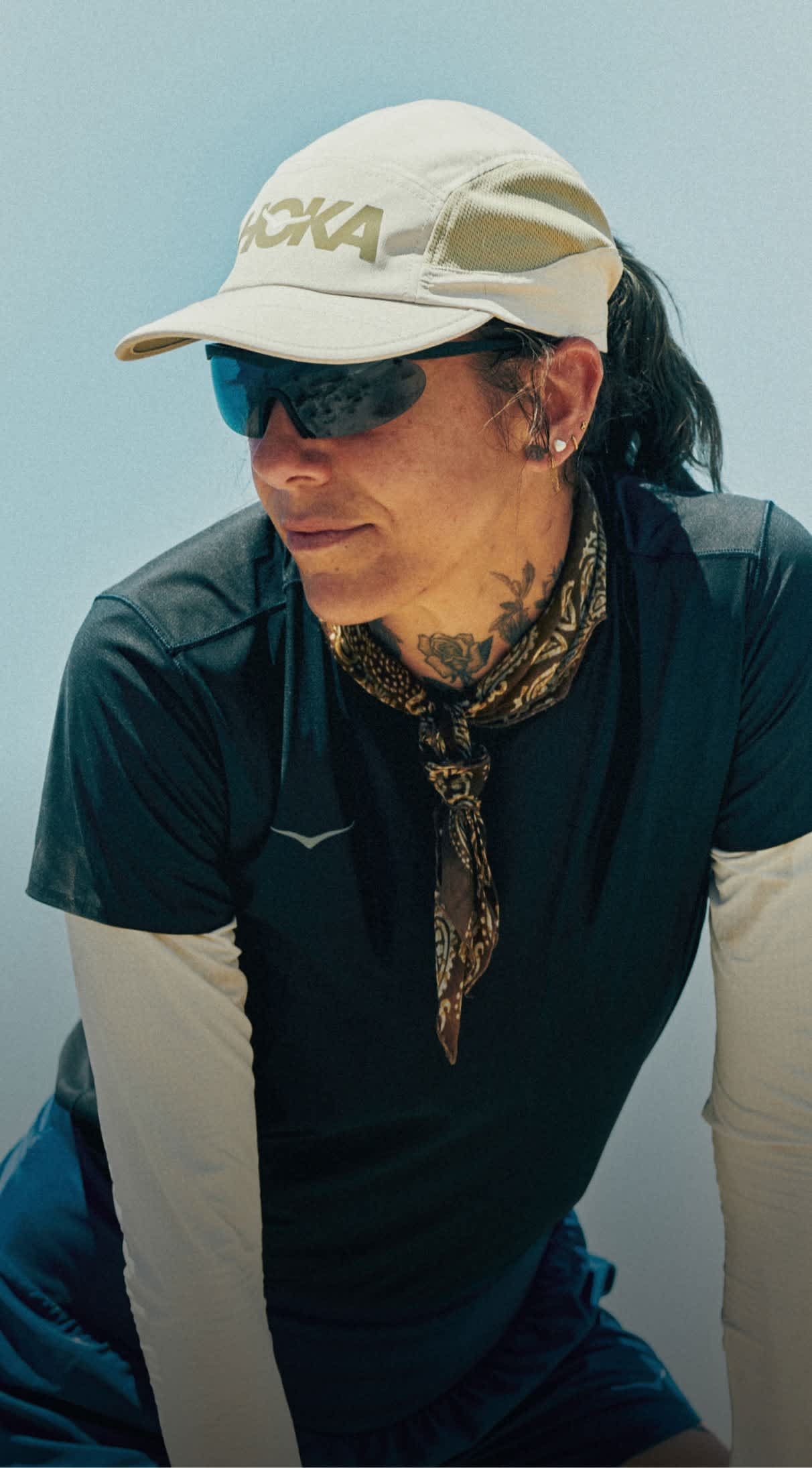 Person in HOKA cap, sunglasses, bandana, and dark shirt; clear blue sky.