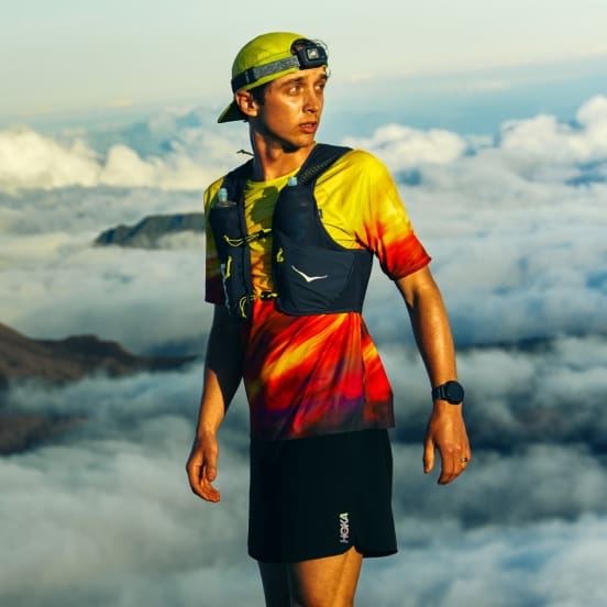 Runner in tie-dye shirt, vest with bottles, cap, headlamp, overlooking mountains.