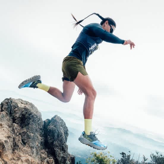 Person in Hoka trail running shoes jumping from a rocky ledge, mountain view.