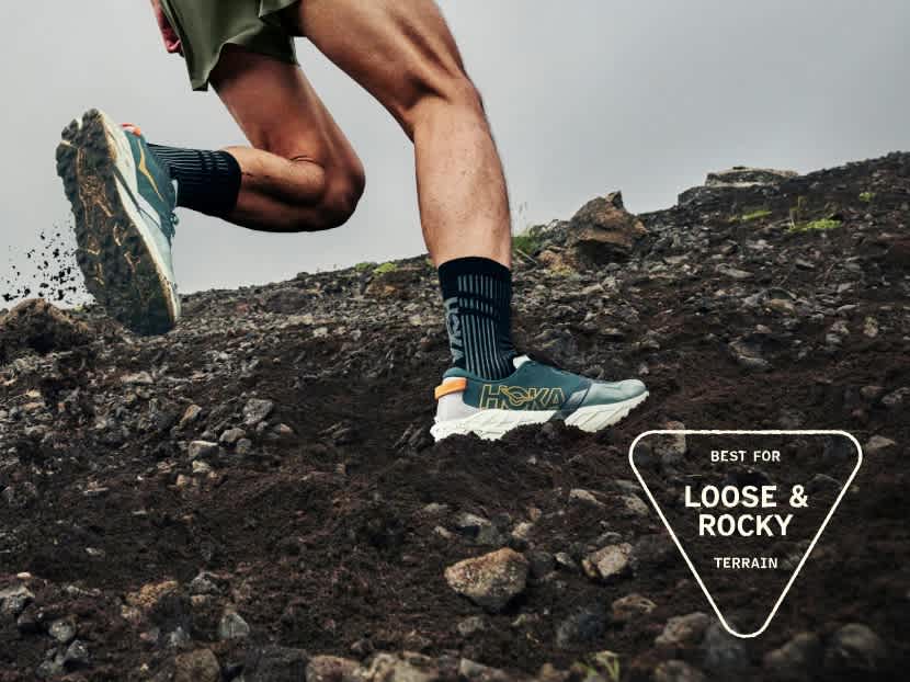 Person running uphill on loose, rocky terrain, dirt flying, showing trail shoes.