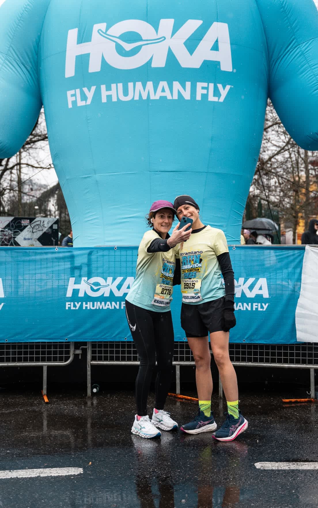 Two individuals in race attire take a selfie at a Hoka-sponsored running event.