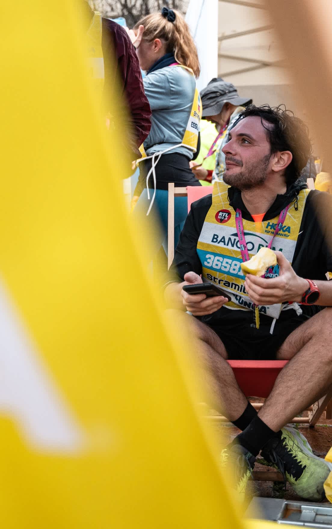 Person in yellow race bib 3658, holding phone and apple, looking right, smiling.