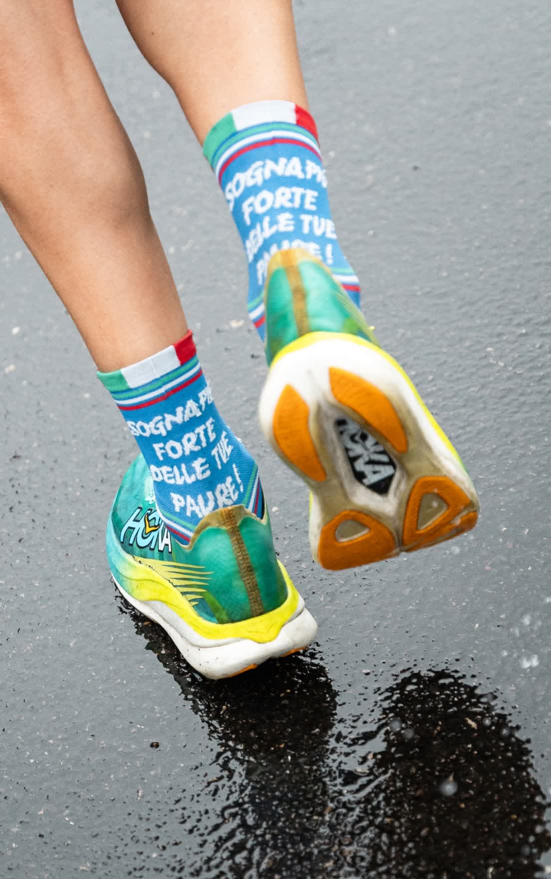 Close-up of runner's feet, HOKA shoes, and 'SOGNAPA FORTE' socks on wet asphalt.