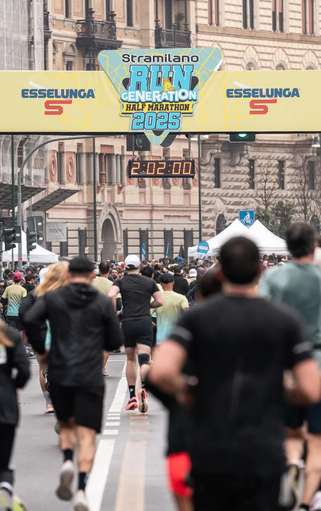 Runners under a 'Stramilano Half Marathon 2025' banner, clock shows 2:02:00.