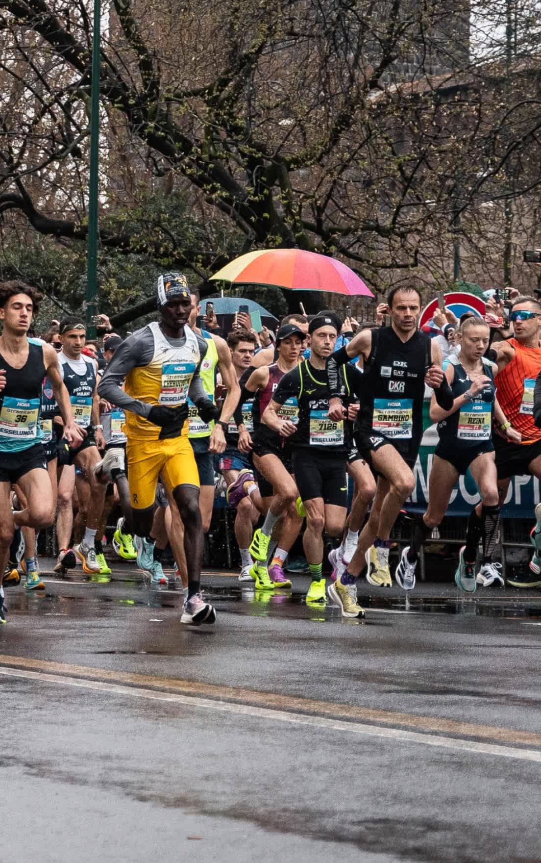 A group of athletes running on a wet street during a race; a colorful umbrella is