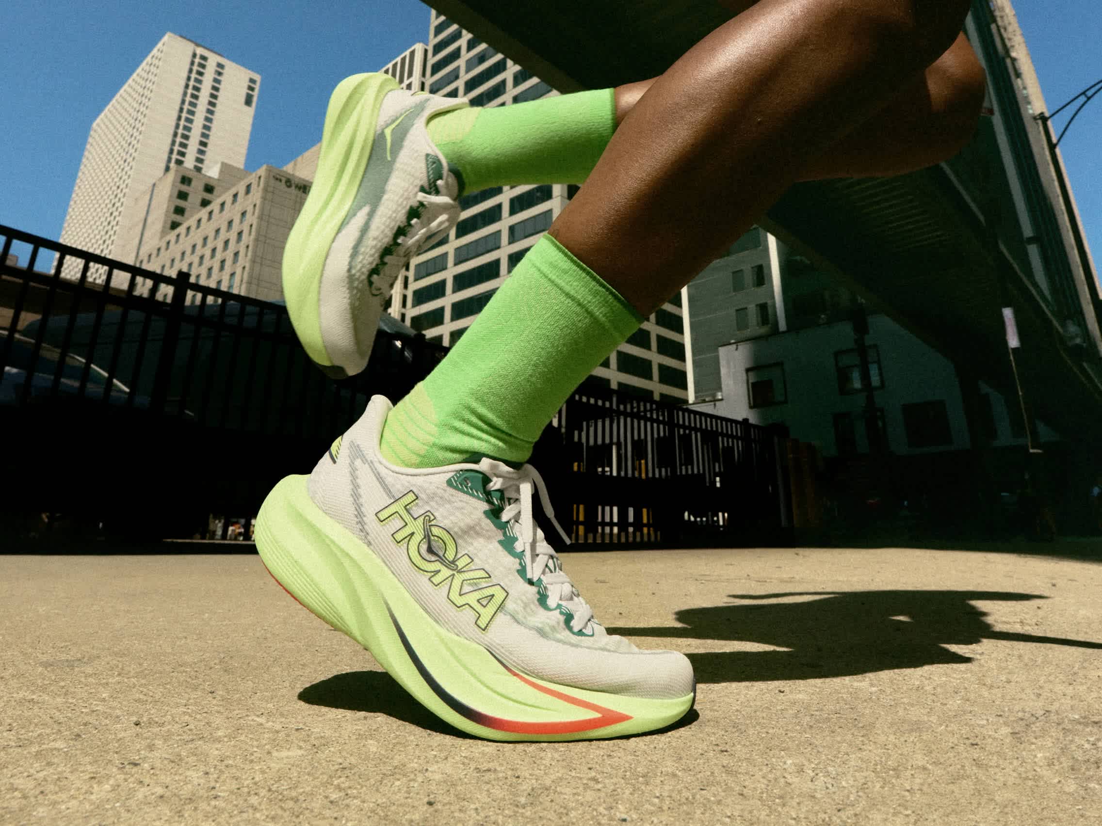Person running in Hoka shoes and neon green socks, urban background with buildings.