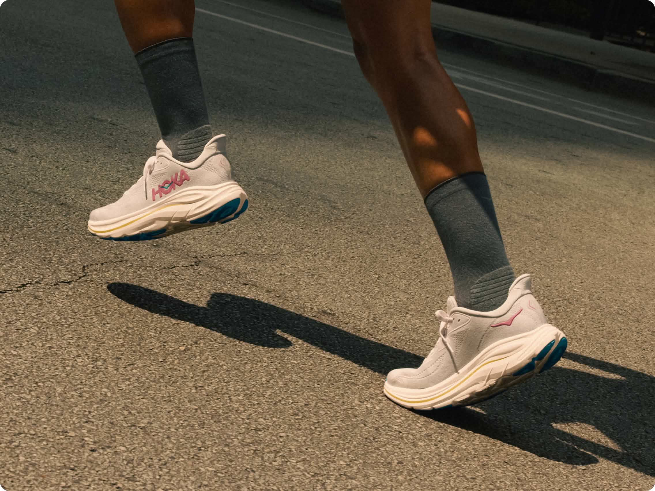 Runner's legs in Hoka shoes on asphalt, one shoe airborne, strong shadows.