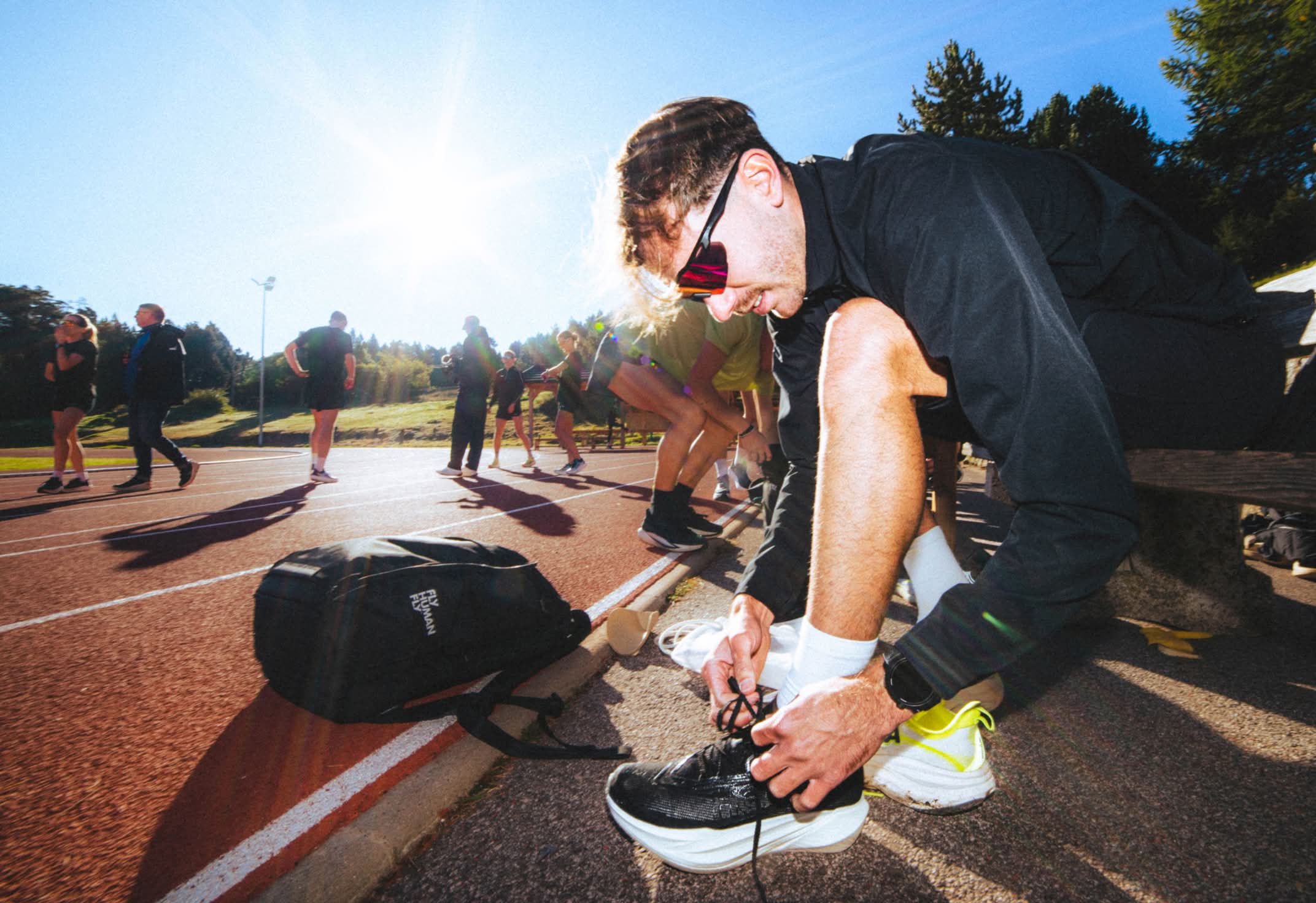 Person tying shoes on a sunny running track with others in the background.