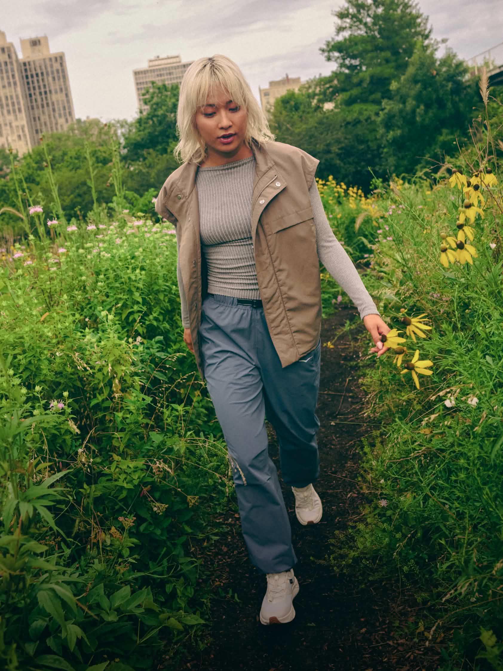 Campaign shot showing a female model wearing the HOKA Transport 2 in a field with urban backdrop.