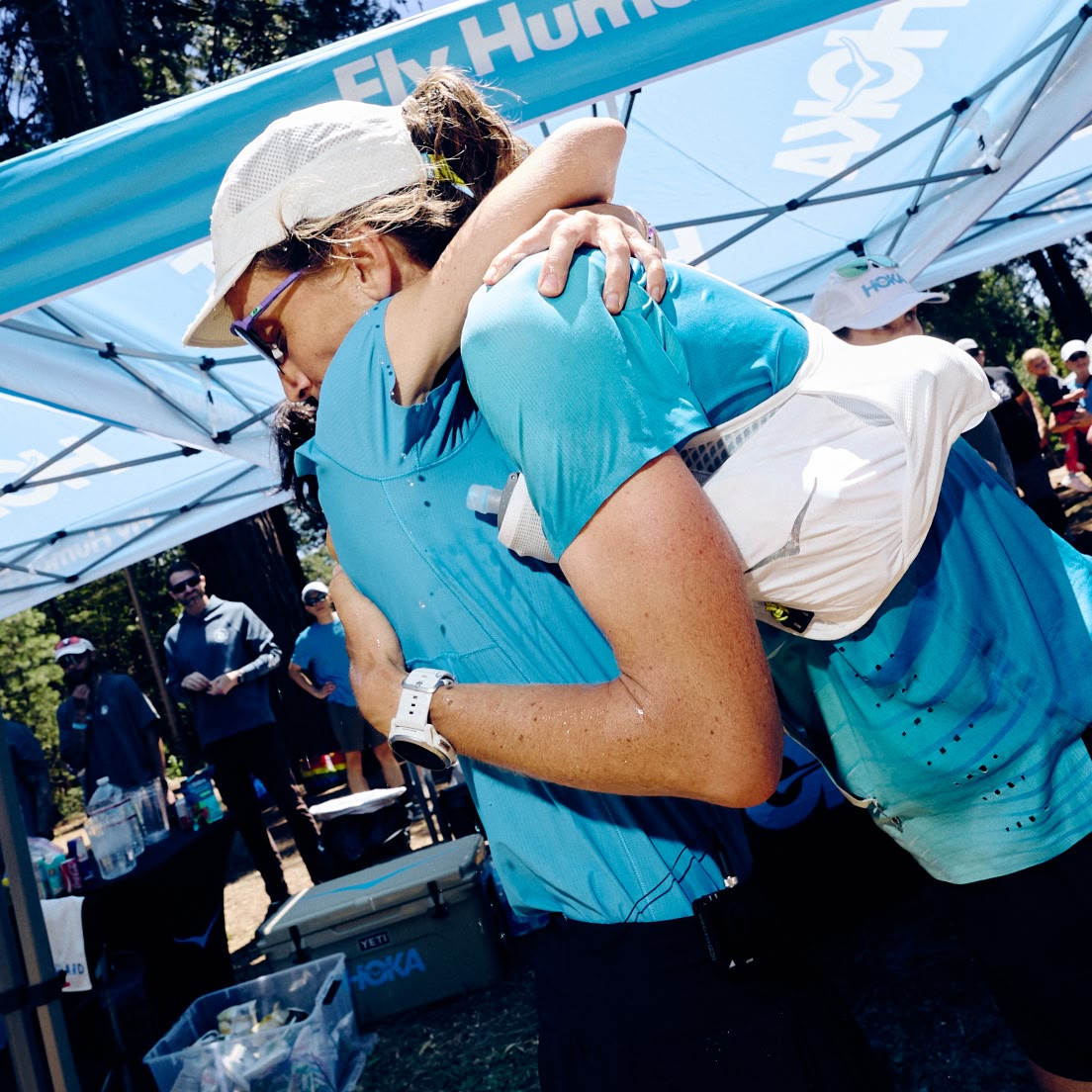 Two individuals embrace near blue HOKA tents after an event.