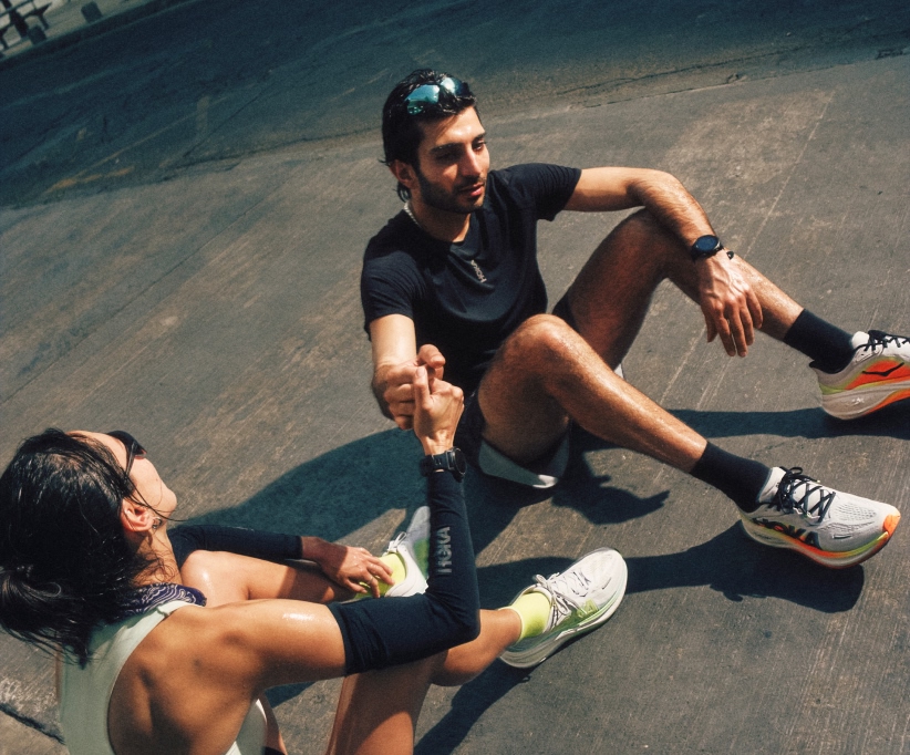 Two individuals sit on pavement, hands clasped, wearing Hoka activewear and shoes.
