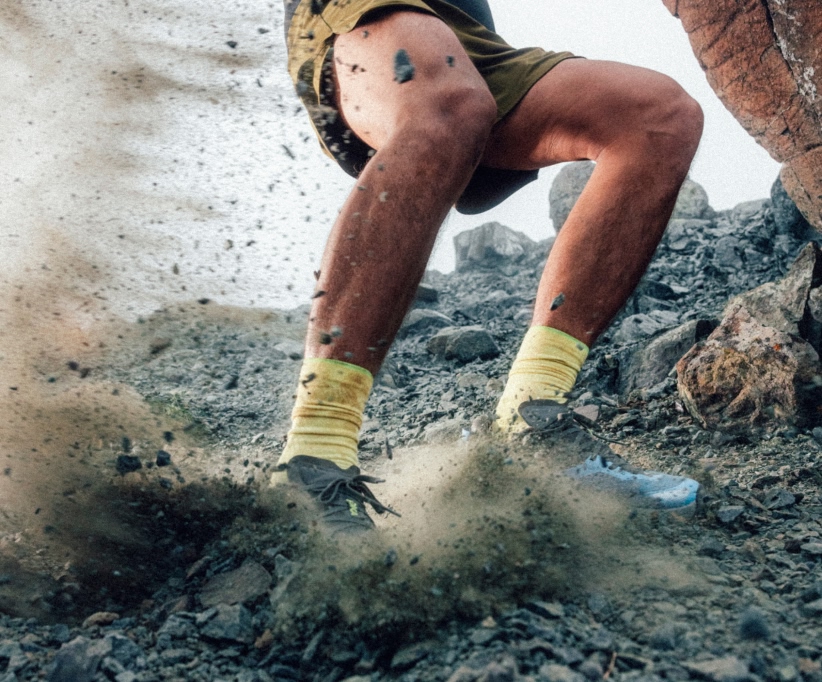 Person's legs in shorts & yellow socks, kicking up dust on rocky terrain.