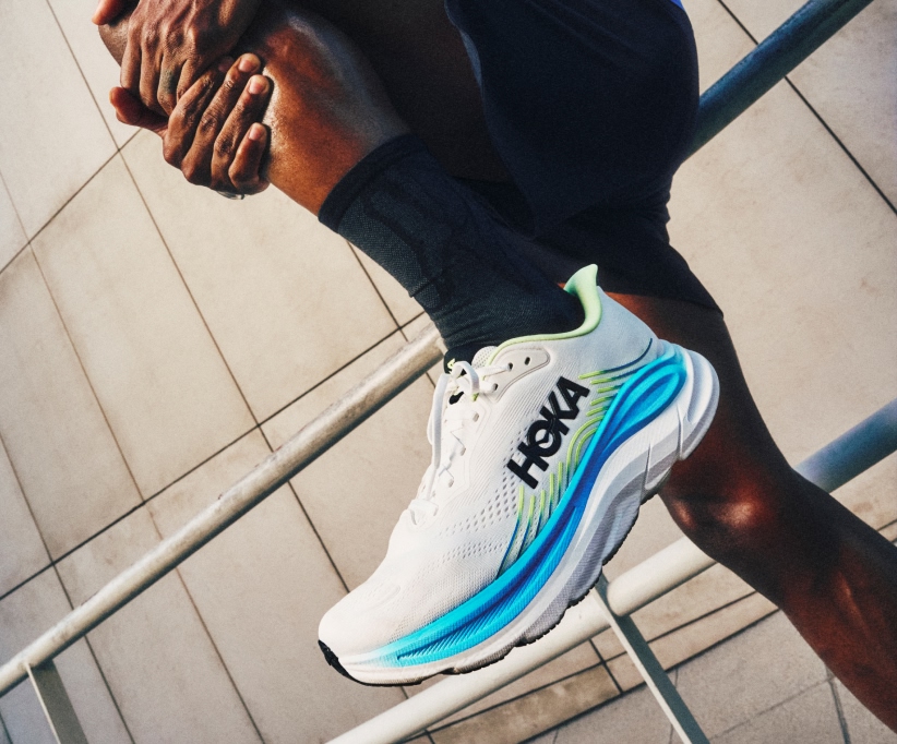 Person stretching leg, wearing white Hoka running shoe with blue and yellow sole