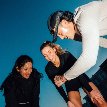 Three individuals in activewear, two smiling down, one leaning, against blue sky.