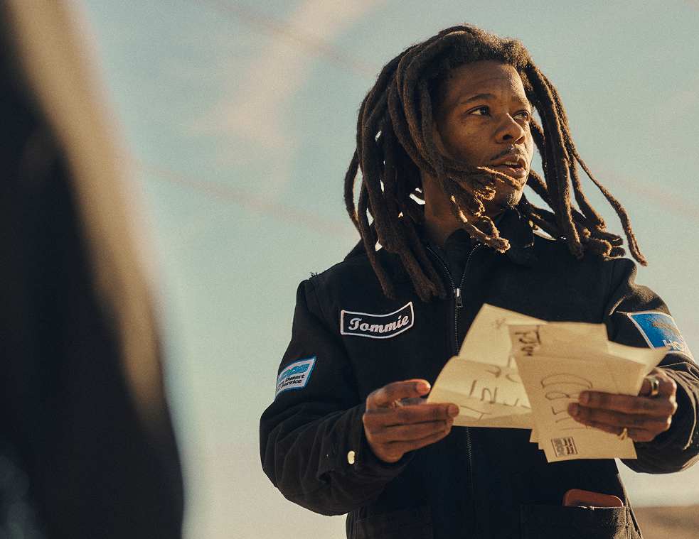 A man with long dreadlocks wearing a black work jacket with a name patch, holding several envelopes and looking off-camera.