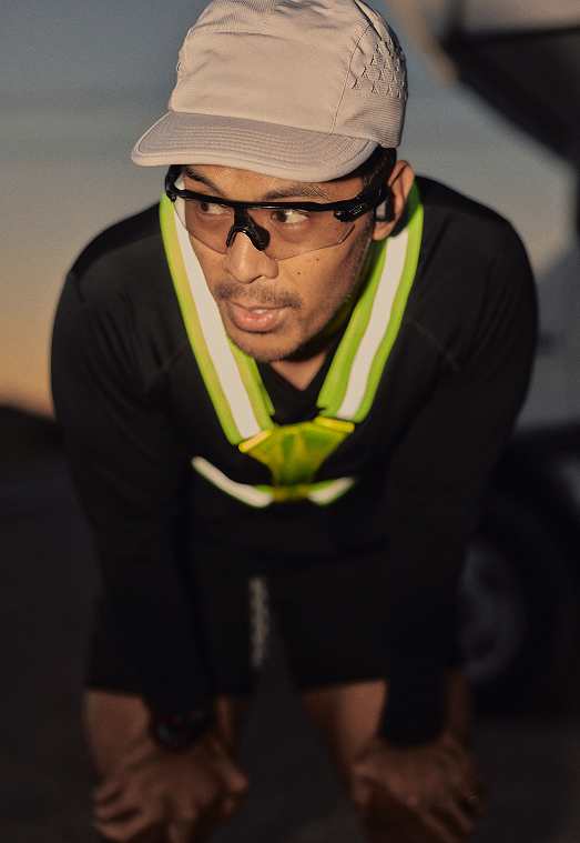 A close-up of a runner wearing a white cap, clear sports glasses, and a neon yellow reflective safety harness, looking focused.