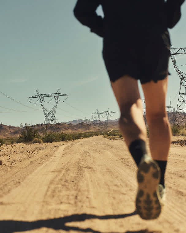 A low-angle, out-of-focus shot from behind a runner on a dirt desert road, with large power line towers stretching into the distance