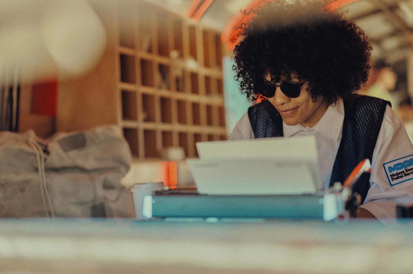 A person with sunglasses types on a vintage typewriter. They wear a "Mojave Desert Postal Service" uniform patch and a dark mesh vest. The setting has a warm, nostalgic aesthetic with blurred wooden shelving in the background.
