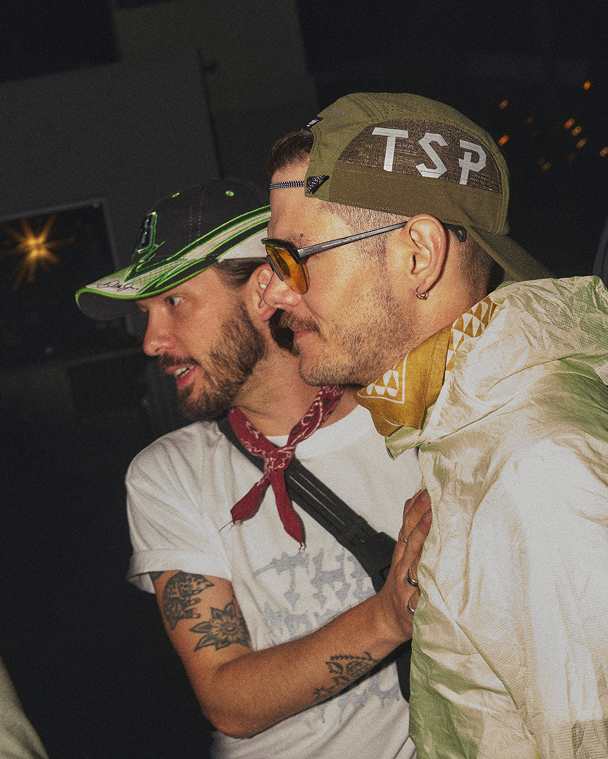Two men standing together at night; one wears an olive "TSP" hat and sunglasses, the other wears a black and green cap and a red bandana.