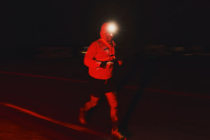 A runner captured in motion at night, illuminated by a bright headlamp and wearing a reflective orange jacket.